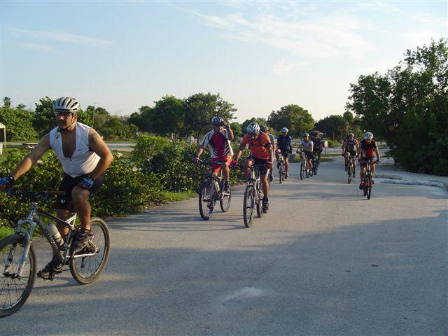 A group of cyclists riding mountain bikes along a paved path surrounded by greenery on a sunny day. The scene features various cyclists wearing helmets and cycling attire, with some riding in the foreground and others in the background. The sky is clear, indicating a bright and pleasant atmosphere for the outdoor activity. Oleta River State Park mountain bike trail.