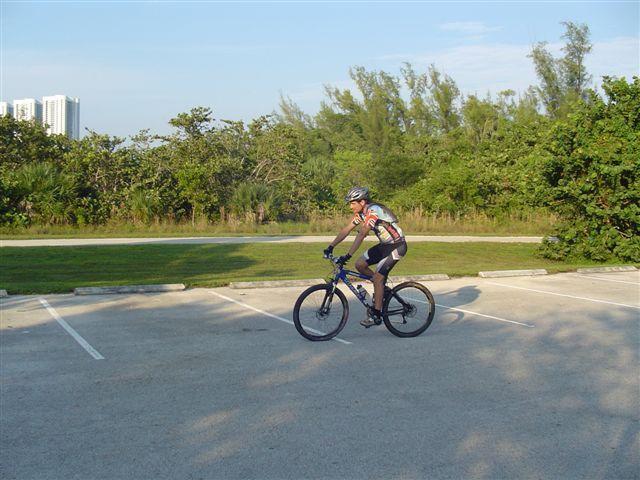 A person riding a mountain bike in a parking lot surrounded by greenery, with tall buildings visible in the background and a clear blue sky above. Oleta River State Park mountain bike trail.