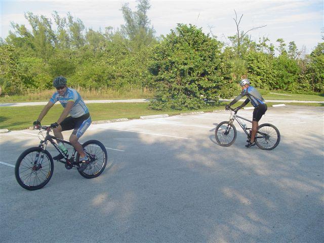 Two cyclists riding their mountain bikes in a parking area surrounded by greenery. One cyclist is in the foreground, while the other rides in the background, both wearing helmets and athletic clothing suitable for biking. Oleta River State Park mountain bike trail.