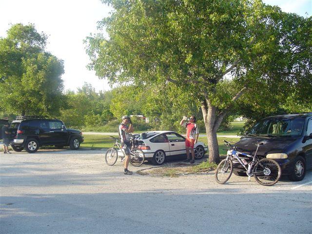 Two cyclists in casual attire stand by a parked car in a gravel lot surrounded by trees. One cyclist is holding their bike, while the other gestures as they converse. Several vehicles, including a minivan and an SUV, are parked nearby. The scene captures a sunny day in a natural setting. Oleta River State Park mountain bike trail.