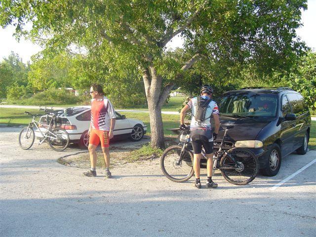 Two cyclists in a parking area preparing for a ride. One cyclist, wearing a red and gray outfit, is walking near a tree, while the other, dressed in a black and red outfit, is adjusting their bike. A white car and a minivan are parked nearby, set against a backdrop of greenery. Oleta River State Park mountain bike trail.