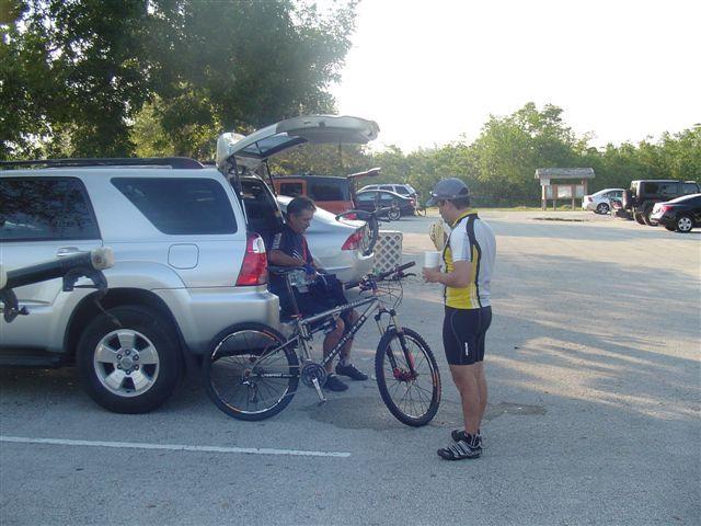Two individuals are interacting in a parking lot. One person is seated on a bicycle, while the other stands beside them, holding a drink. A silver SUV with its trunk open is in the background, and several cars are parked nearby. The scene is set in a natural environment with trees in the background, indicating an outdoor activity such as biking. Oleta River State Park mountain bike trail.