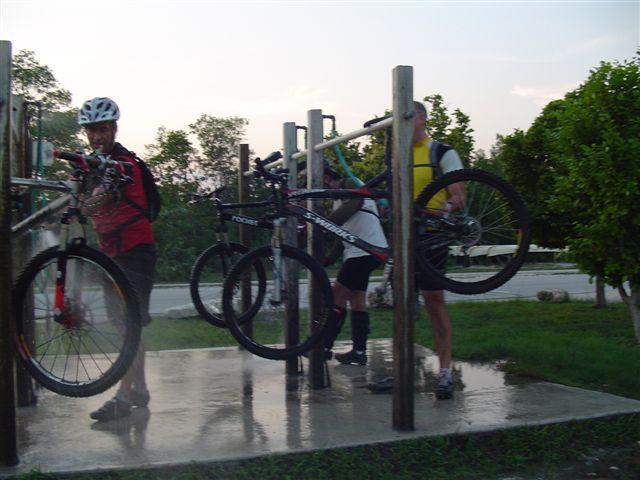 Two cyclists are washing their mountain bikes at a bike cleaning station, surrounded by greenery and a paved area. One cyclist is in a red jersey, handling a bike, while the other, wearing a yellow flotation device, is lifting a bike onto the cleaning rack. The scene is set during dusk, with a road visible in the background. Oleta River State Park mountain bike trail.