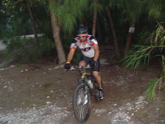A mountain biker riding a bicycle along a dirt trail surrounded by trees. The cyclist is wearing a helmet and a short-sleeved jersey, appearing focused as he navigates the uneven terrain. The scene is set in a wooded area with greenery in the background. Oleta River State Park mountain bike trail.