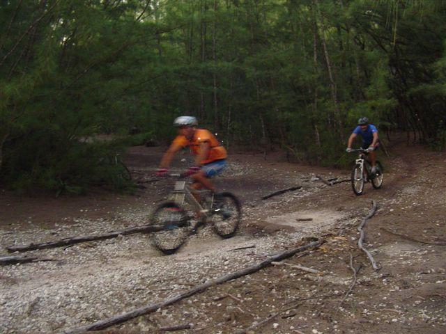 Two mountain bikers riding along a dirt trail through a densely wooded area, with trees and scattered branches in the background. One rider is wearing an orange shirt and the other is in a blue shirt. The image captures a sense of movement as they navigate the uneven terrain. Oleta River State Park mountain bike trail.