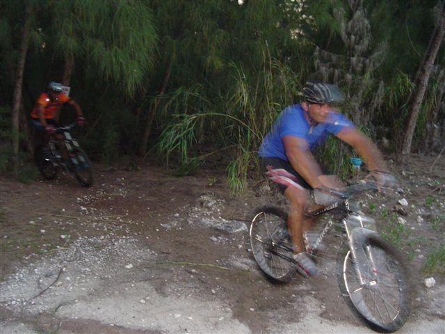 Two mountain bikers navigate a dirt trail surrounded by trees. One cyclist, wearing a blue shirt and helmet, is prominently in motion, while the other, clad in an orange shirt, trails behind. The image conveys a sense of speed and adventure in a natural setting. Oleta River State Park mountain bike trail.