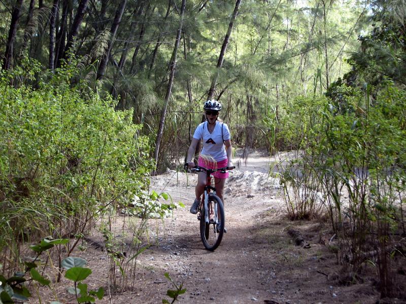 A person riding a mountain bike along a dirt trail surrounded by tall trees and lush greenery. The cyclist is wearing a helmet and a white shirt with colored shorts, demonstrating an outdoor recreational activity. Oleta River State Park mountain bike trail.