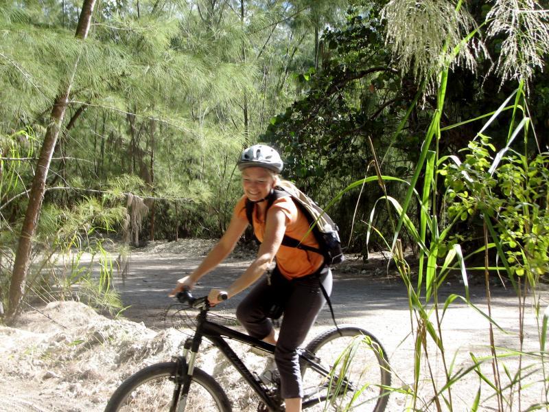 A person riding a black mountain bike on a dirt path surrounded by greenery, including trees and tall grass. They are wearing an orange shirt and a helmet, smiling as they enjoy their ride in a natural setting. Oleta River State Park mountain bike trail.