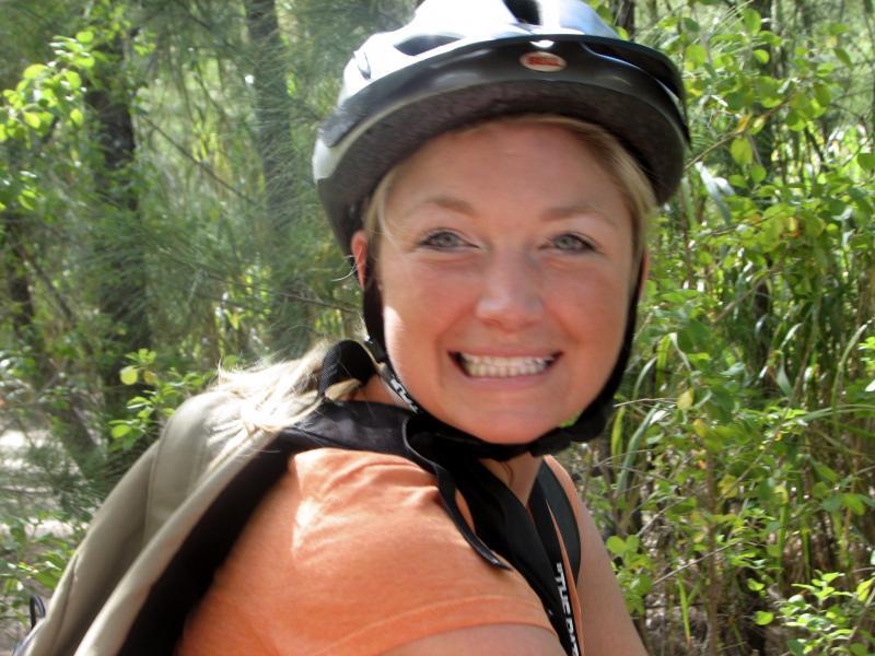 A smiling woman wearing a bicycle helmet and an orange shirt, posing in a green, wooded area. Oleta River State Park mountain bike trail.