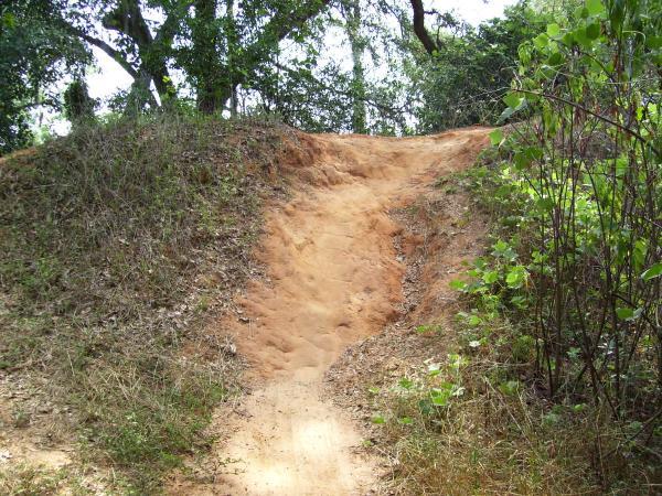 A sloping dirt path leading up to a raised mound, surrounded by sparse vegetation and trees. The terrain appears dry, with patches of grass and light shrubbery on either side. Tom Brown / Lafayette Heritage Park mountain bike trail.