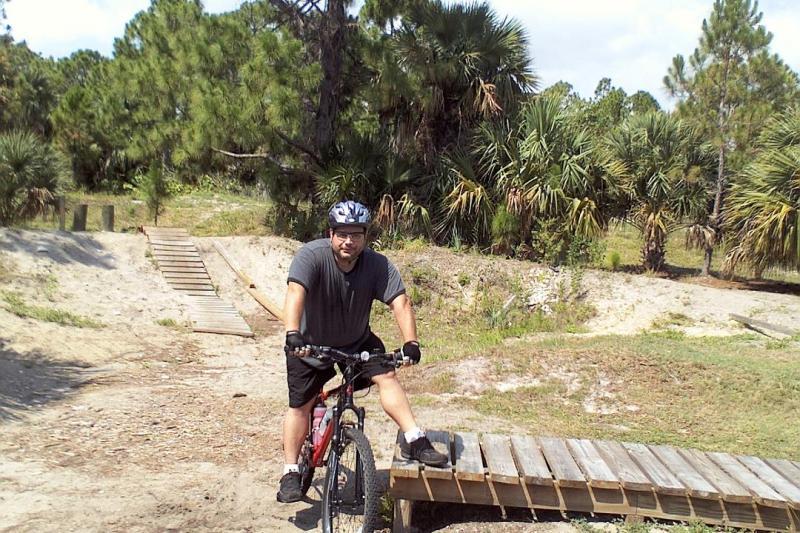 A person wearing a helmet is riding a bicycle and standing on a wooden ramp in a natural outdoor setting with lush greenery and palm trees in the background. The individual appears focused and is positioned near a dirt trail that leads deeper into the landscape. Jonathan Dickinson State Park mountain bike trail.