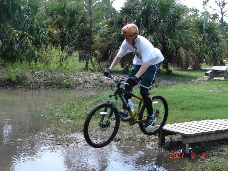 A person wearing a helmet and protective gear jumps off a wooden platform while riding a mountain bike over a small puddle in a natural setting, surrounded by greenery. Jonathan Dickinson State Park mountain bike trail.