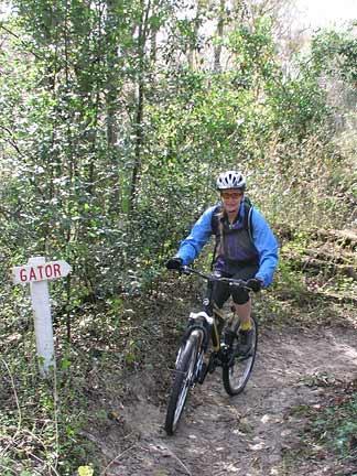 A person riding a mountain bike on a dirt trail surrounded by lush greenery, with a sign labeled "GATOR" visible in the foreground. The cyclist is wearing a helmet and a blue jacket. Morris Bridge Park mountain bike trail.