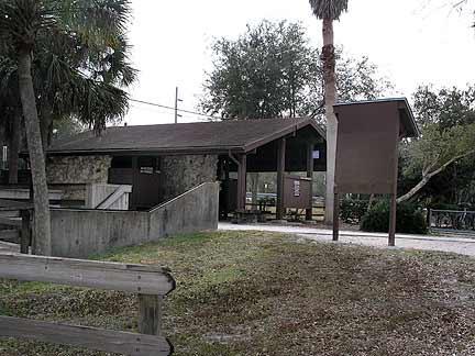A small, rustic building with a wooden exterior surrounded by greenery and palm trees. There is a signboard near the entrance and a slope leading to a shaded area. The scene is set in a natural, outdoor environment. Morris Bridge Park mountain bike trail.