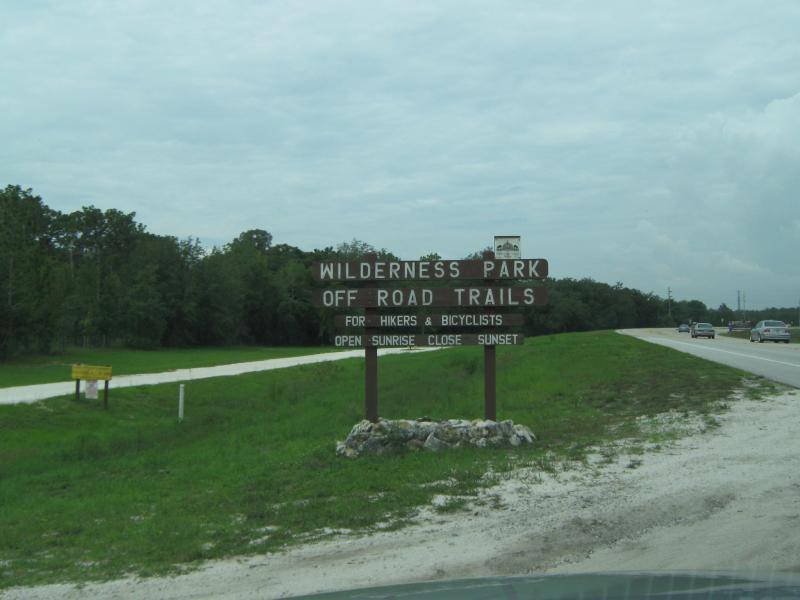Signpost for Wilderness Park Off Road Trails, indicating it's open for hikers and cyclists from sunrise to sunset, with a view of a grassy area and nearby road. Morris Bridge Park mountain bike trail.