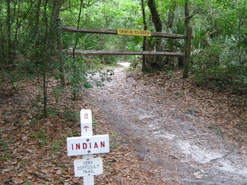 A wooded trail entrance with a closed gate and a sign reading "TRAILS CLOSED" above it. In front of the gate, a white sign indicates "INDIAN" with an arrow pointing forward and the words "VERY DIFFICULT TRAIL." The path ahead is partially visible, surrounded by lush greenery and fallen leaves. Morris Bridge Park mountain bike trail.