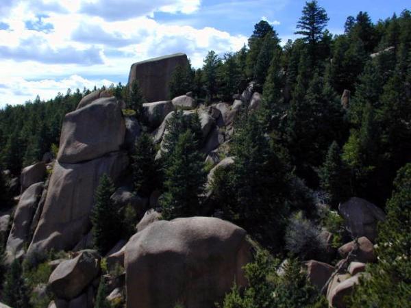 Rocky hillside with large boulders and dense evergreen trees under a partly cloudy sky. Monument Preserve mountain bike trail.