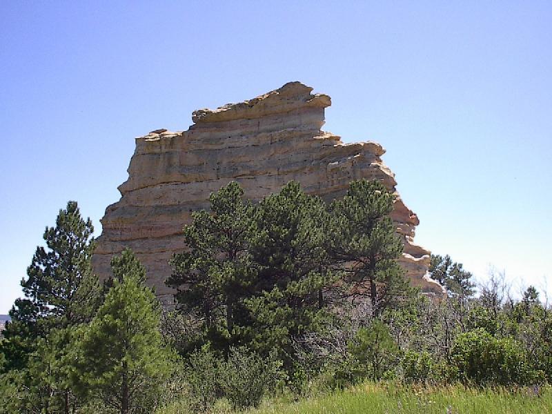 A towering rock formation rises prominently against a clear blue sky, surrounded by dense green trees and shrubs. The unique geological structure features layered rock with a flat top, creating a dramatic landscape in a natural setting. Monument Preserve mountain bike trail.