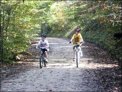 Two children riding bicycles on a dirt path surrounded by trees, with fallen leaves covering the ground. One child wears a white shirt and helmet, while the other wears a yellow shirt and helmet. The scene captures a sunny day in a forested area. Salem Lake mountain bike trail.