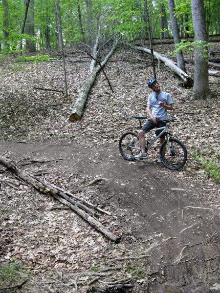 A young man wearing a blue helmet and casual clothing stands beside a mountain bike on a dirt path in a wooded area. He is looking up with a thoughtful expression, surrounded by trees and fallen logs, with scattered leaves covering the ground. The scene captures a moment of contemplation in a natural outdoor setting. Schaeffer Farms mountain bike trail.
