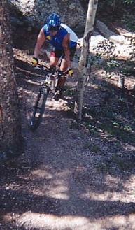 A person wearing a blue helmet and a sleeveless blue and white jersey is riding a mountain bike down a narrow, wooded trail surrounded by trees. The terrain is uneven, with patches of dirt and gravel. Rampart Reservoir mountain bike trail.
