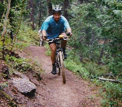 A person riding a mountain bike along a narrow dirt trail in a forested area. The cyclist is wearing a helmet and a blue tie-dye shirt, surrounded by greenery and trees. Rampart Reservoir mountain bike trail.