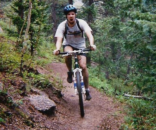 A man enthusiastically riding a mountain bike along a dirt trail, surrounded by greenery and trees. He is wearing a helmet and a T-shirt, and appears to be enjoying the ride. Rampart Reservoir mountain bike trail.