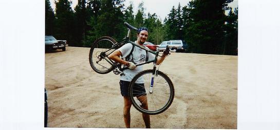A smiling person wearing a bandana and casual clothing is standing in a gravel parking area, holding a mountain bike over their shoulder with one hand and the bike's front wheel in the other. In the background, there are several parked cars and a forested area. Rampart Reservoir mountain bike trail.