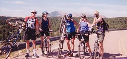 A group of five mountain bikers standing on the side of a road surrounded by trees and mountains. They are posing with their bikes, smiling, and wearing helmets and athletic clothing. The sunny sky and scenic landscape create a cheerful outdoor atmosphere. Rampart Reservoir mountain bike trail.