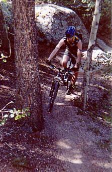 A person riding a mountain bike on a narrow trail surrounded by trees and large rocks in a forested area. Rampart Reservoir mountain bike trail.