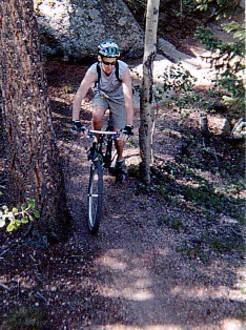 A person riding a mountain bike on a narrow trail surrounded by trees and rocks, wearing a helmet and athletic clothing. The scene captures a natural outdoor setting, showcasing the rugged terrain ideal for biking. Rampart Reservoir mountain bike trail.