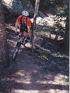 A mountain biker in an orange shirt and helmet descending a wooded trail, surrounded by trees and rocky terrain. Sunlight filters through the branches, casting shadows on the path. Rampart Reservoir mountain bike trail.