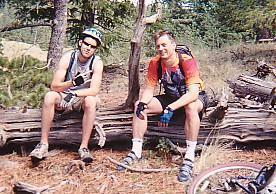 Two men taking a break while mountain biking in a wooded area. One man is sitting on a log, wearing a helmet and a tank top, giving a thumbs-up. The other man, sitting alongside him, is wearing a colorful shirt and gloves, also posing for the camera. In the foreground, a mountain bike is partially visible. Rampart Reservoir mountain bike trail.