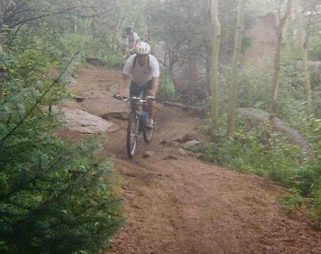 A mountain biker riding down a dirt trail surrounded by lush greenery and trees, with rocky terrain visible in the background. Rampart Reservoir mountain bike trail.