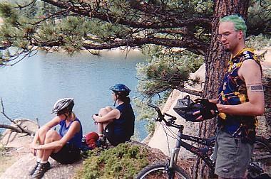 A group of three cyclists enjoying a break by a serene lake surrounded by trees. Two individuals sit on a rock, one wearing a helmet and casual cycling attire, while another stands nearby with green hair, holding a cycling glove. A mountain bike is positioned next to the standing cyclist. Rampart Reservoir mountain bike trail.