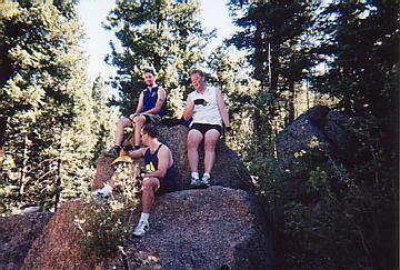 Three people sitting on a large rock in a forested area, surrounded by tall trees. They are dressed in casual athletic clothing and appear to be enjoying a break during an outdoor activity. Rampart Reservoir mountain bike trail.