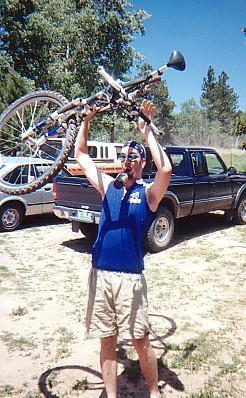 A man wearing a blue tank top and shorts is joyfully lifting a bicycle over his head in an outdoor setting, with trees and parked vehicles in the background. Rampart Reservoir mountain bike trail.