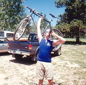 A person in a sleeveless blue shirt and shorts stands outside, lifting a bicycle above their head with one hand while drinking from a can with the other. A pickup truck is parked in the background, surrounded by trees. Rampart Reservoir mountain bike trail.