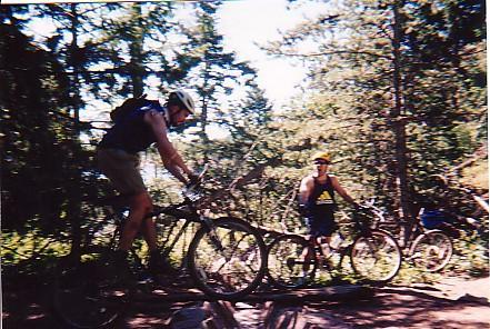 Two mountain bikers riding on a forest trail. One rider is in mid-jump over a log, while the other watches from the side. The scene is surrounded by trees and greenery, indicating a sunny outdoor setting. Rampart Reservoir mountain bike trail.