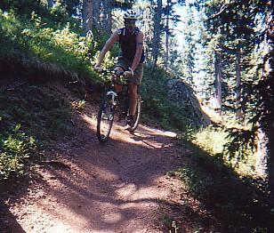 A mountain biker riding on a dirt trail through a forest, surrounded by tall trees and dappled sunlight. The biker is wearing a helmet and shorts, navigating a winding path. Rampart Reservoir mountain bike trail.