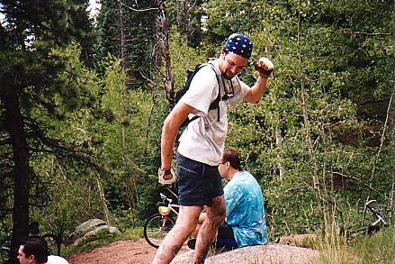 A man wearing a bandana and casual hiking clothes stands on a rock in a forested area, raising his arm as if celebrating or showing off a small object. In the background, a bicycle is visible, and two other people sit nearby, slightly out of focus. The scene is surrounded by green trees and natural foliage. Rampart Reservoir mountain bike trail.
