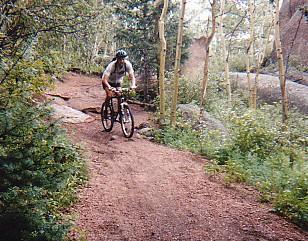 A person riding a mountain bike on a narrow dirt trail surrounded by trees and rocks in a natural outdoor setting. Rampart Reservoir mountain bike trail.