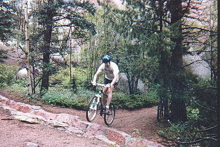 A person riding a mountain bike on a narrow trail surrounded by dense trees and greenery. The cyclist is wearing a helmet and appears to be navigating a rocky section of the path. Rampart Reservoir mountain bike trail.
