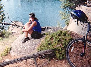A woman wearing a helmet sits on a large rock by a body of water, smiling at the camera. She is dressed in a blue tank top and black shorts, and a mountain bike with a helmet resting on it is visible nearby. The scene is surrounded by trees and features a serene, natural landscape. Rampart Reservoir mountain bike trail.