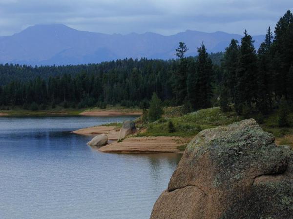 A tranquil landscape depicting a calm lake surrounded by trees and rocky shores, with distant mountains under a cloudy sky. Rampart Reservoir mountain bike trail.