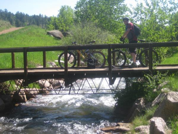 A person stands on a wooden bridge over a flowing stream, with two mountain bikes parked beside them. The background features lush green trees and a trail, suggesting a scenic outdoor location. Sunlight brightens the scene, enhancing the vibrant greenery. Rampart Reservoir mountain bike trail.