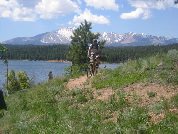 A mountain biker riding along a dirt path near a lake, surrounded by green vegetation and trees, with snow-capped mountains in the background under a partly cloudy sky. Rampart Reservoir mountain bike trail.