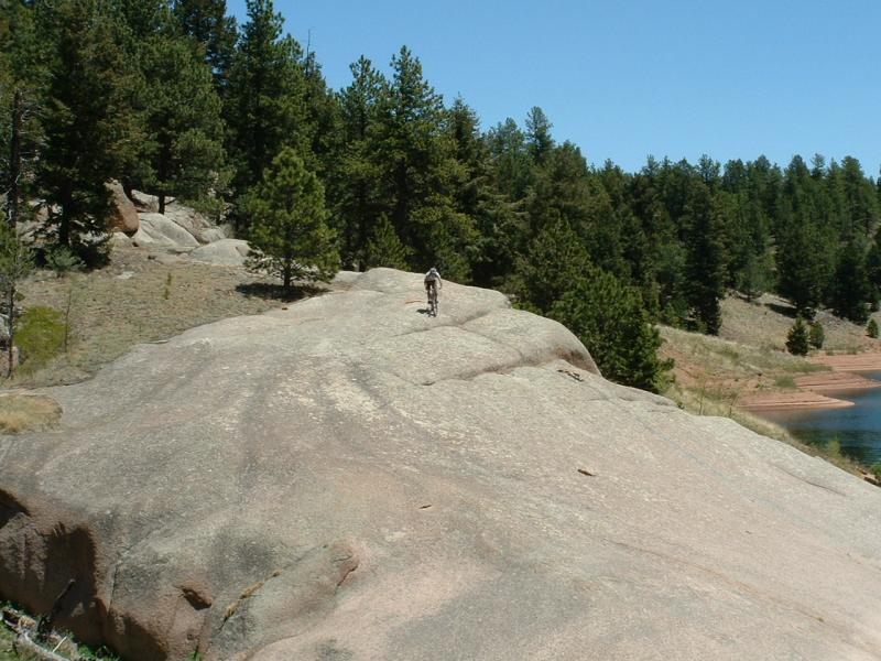 A person in hiking gear is walking along a large, rocky surface with a forest of pine trees in the background. A calm body of water is visible to the right, under a clear blue sky. Rampart Reservoir mountain bike trail.