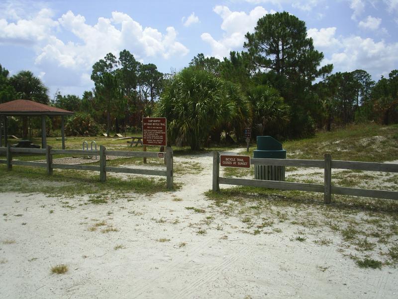 A sandy pathway leading into a wooded area with palm trees and shrubs, flanked by wooden fences. Informational signs about hiking and biking trails are visible, along with a covered gazebo and a trash bin in the background. The sky is partly cloudy, creating a bright outdoor atmosphere. Jonathan Dickinson State Park mountain bike trail.
