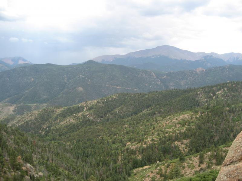 A panoramic view of rolling green mountains under a cloudy sky, with distant peaks emerging in the background. The landscape features a mix of coniferous trees and rugged terrain, creating a serene natural setting. Waldo Canyon mountain bike trail.